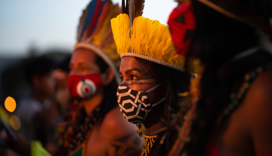 An Indigenous woman wearing a yellow feather headdress and a black and white face stands between two other activists wearing similar attire at a demonstration in Brazil.