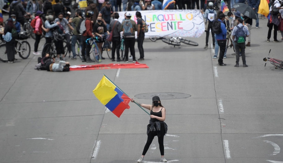 A woman wearing a black face mask and a black tank top and tights waves a Colombian flag in the middle of a large street during a demonstration, while other protester march behind her.