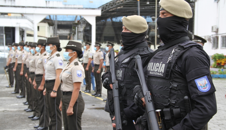 Two police officers carrying heavy guns and wearing and black bulletproof vests and body armour stand beside of row of women police officers wearing dark green slacks and short-sleeve khaki shirts.