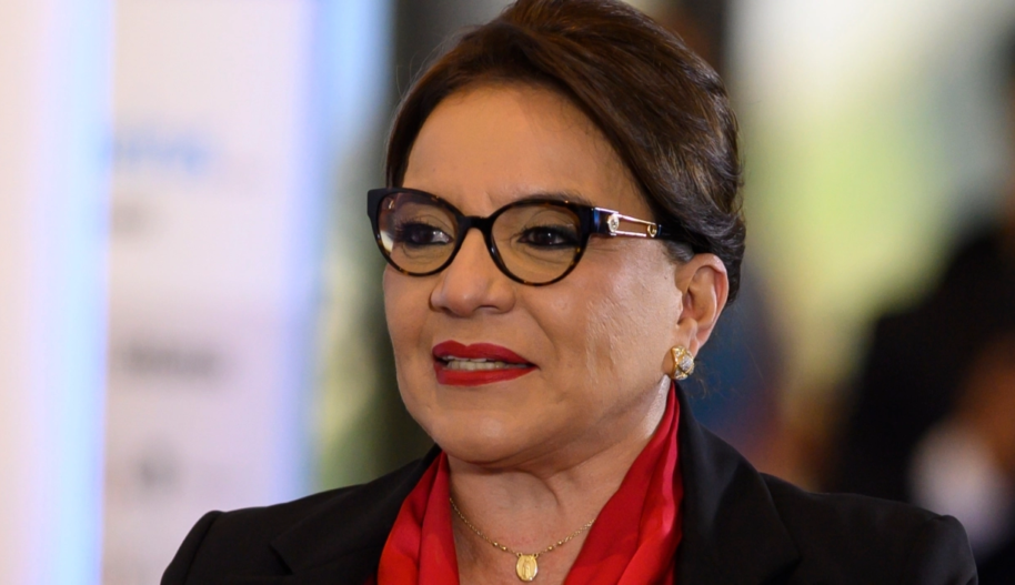 A middle-aged women with black-rimmed cat-eye glasses, brown hair that is parted in the centre, a black blazer, and a red, collared blouse smiles.