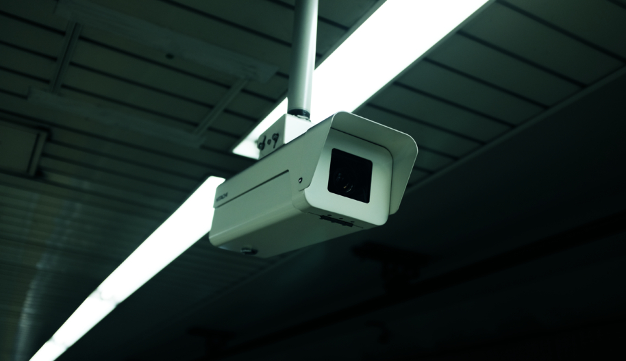 White surveillance camera monitoring a dark space with lined ceiling and overhead lights