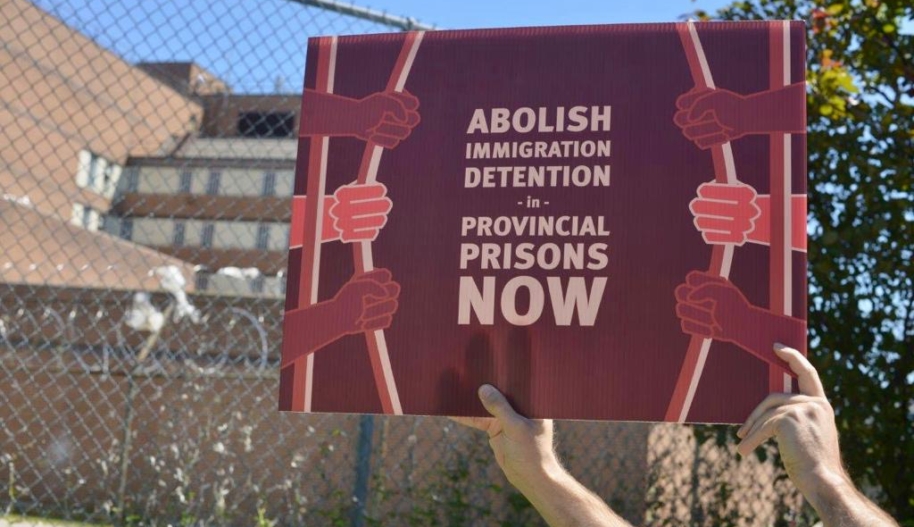 Two hands hold up a sign that reads, "Abolish Immigration Detention in Provincial Jails Now."
