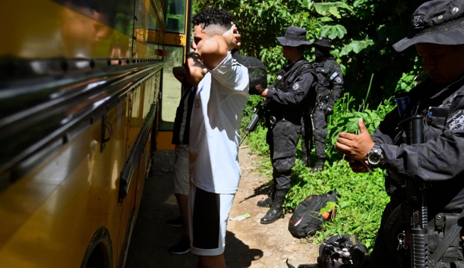 Two men stand in front of a school bus while police in tactical uniforms frisk them.