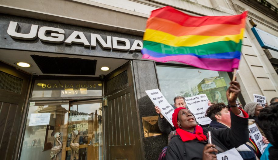 A woman with black jacket, a red had and a red scarf waves a rainbow flag at a protest in in front of a building labelled "Uganda House" on its front door.