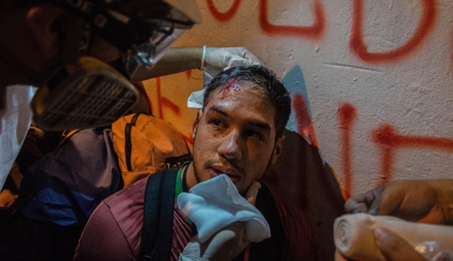 A young man with a wound on his head is leaning against a wall while a medic treats his injury.