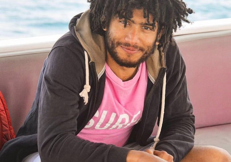 Badr Mohamed seated on a bench wearing a pink t-short and a black cardigan