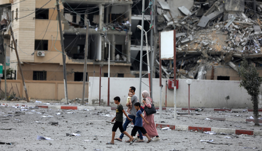 A woman, two boys, and a man carrying a baby walking on a road in front of a bombed-out apartment building.