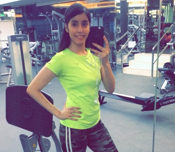 A picture of Manahel wearing a green short sleeve shirt and yoga pants. Her hair is in a ponytail and she appears to have taken the photo in a gym.