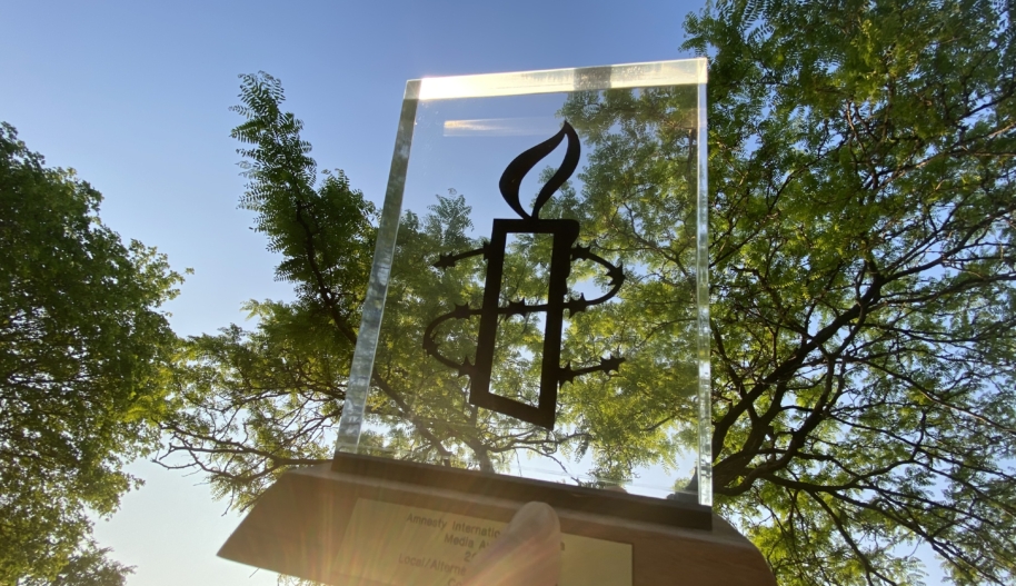 A hand holding a rectangular glass trophy with a brown wooden base under a tree canopy