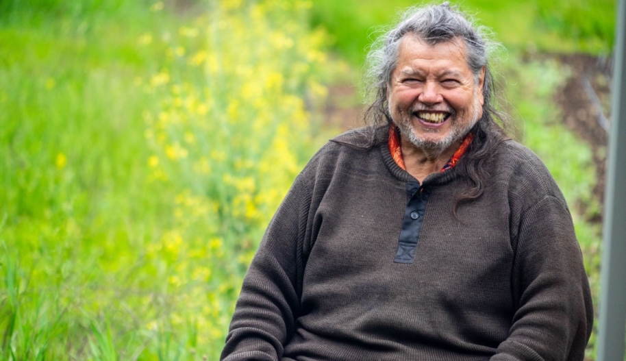 A man in a dark brown and long silver hair smiles while seated.